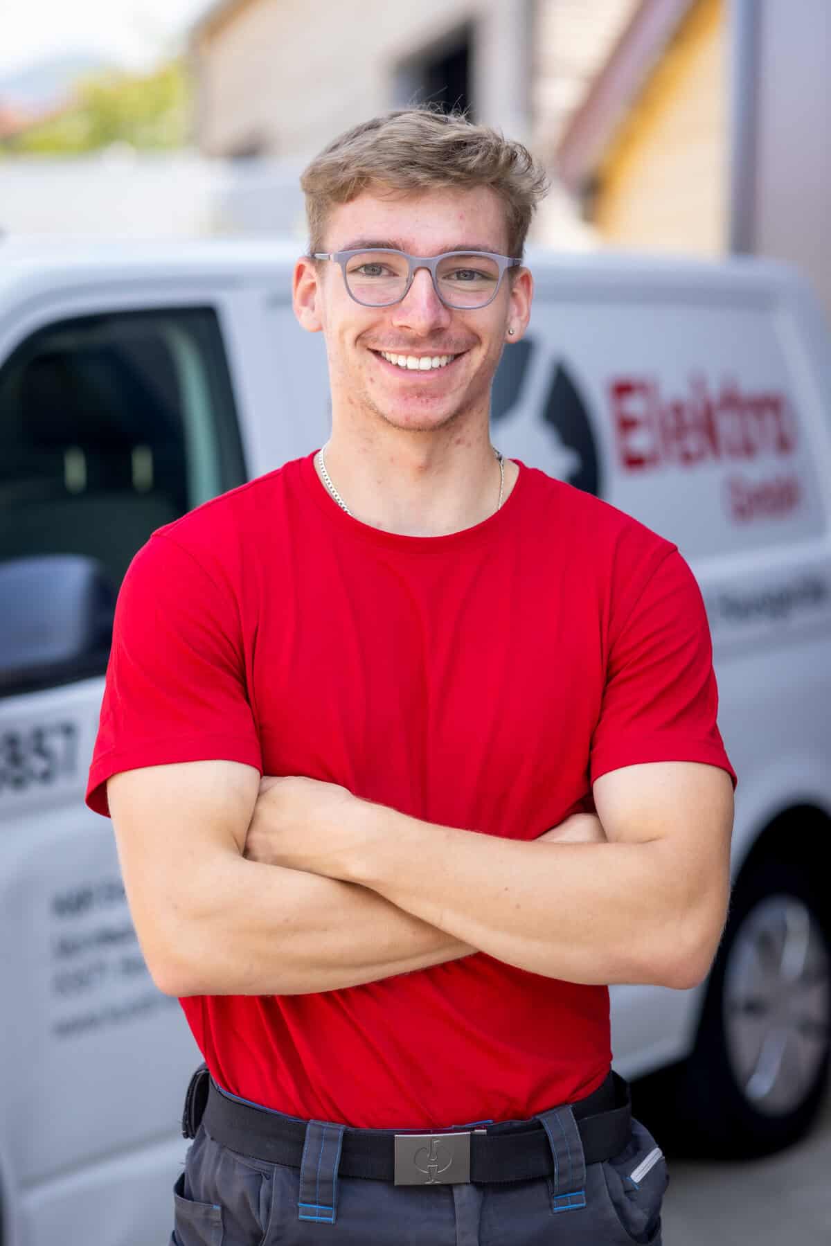 Lächelnder Elektriker in rotem T-Shirt vor Firmenfahrzeug.